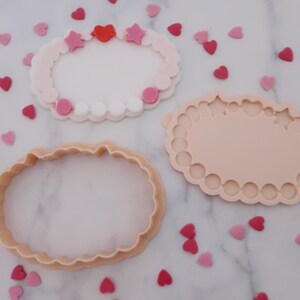 May include: Three cookie cutters in the shape of an oval with a scalloped edge. One is a cutter, one is a mold, and the third is a decorated cookie with pink and red heart and star sprinkles. The background is white with scattered heart sprinkles.