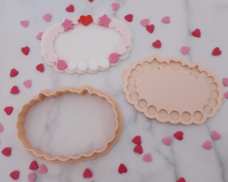 May include: Three cookie cutters in the shape of an oval with a scalloped edge. One is a cutter, one is a mold, and the third is a decorated cookie with pink and red heart and star sprinkles. The background is white with scattered heart sprinkles.
