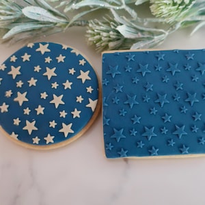May include: Two decorated biscuits, one circular and one square, featuring a deep blue fondant topping. The circular biscuit has white star-shaped cutouts, while the square biscuit features embossed blue stars across its entire surface.