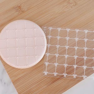 May include: A light pink, circular coaster and a clear, square coaster featuring a grid pattern of white bows and dotted lines. The coasters are on a wooden surface.