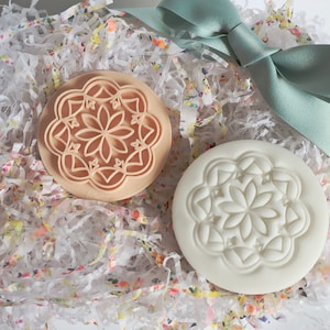 May include: Two round cookie stamps with a floral design. One stamp is beige and raised, the other is white and pressed. The stamps are on a bed of white shredded paper with a floral pattern and a pale green ribbon.