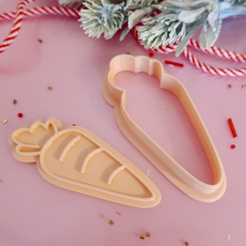 May include: Two light peach carrot-shaped cookie cutters. One is a solid shape, the other an outline. They are on a pink surface with decorative elements.