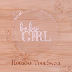 May include: A clear, circular acrylic cake topper with the words "baby GIRL" in a delicate, cursive font. The topper has a small heart-shaped detail with the letters "2TOH". The background is a light-colored wooden surface with the text "House of Toot Sweet".