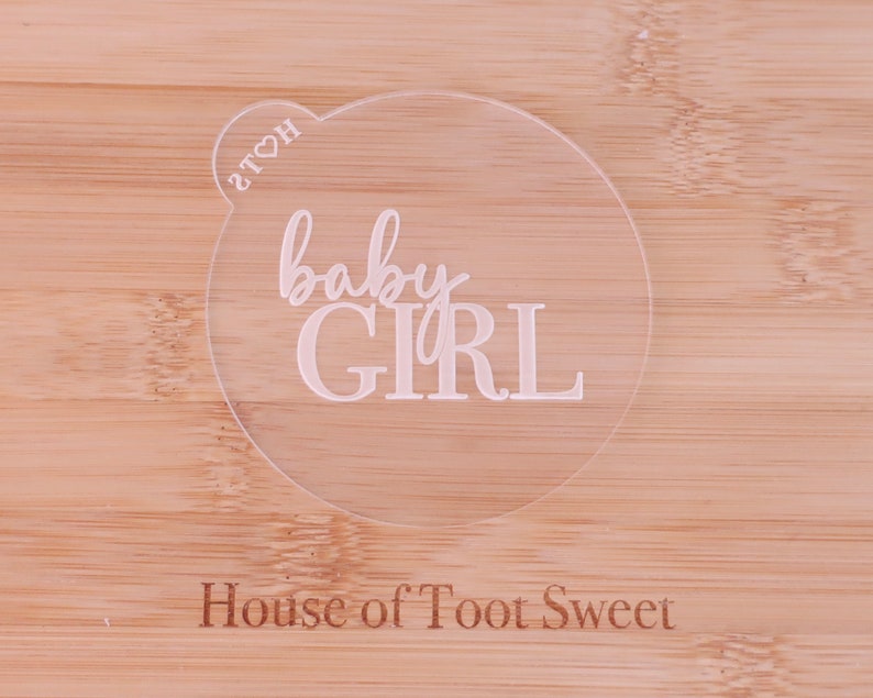 May include: A clear, circular acrylic cake topper with the words "baby GIRL" in a delicate, cursive font. The topper has a small heart-shaped detail with the letters "2TOH". The background is a light-colored wooden surface with the text "House of Toot Sweet".