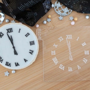 May include: An image featuring a frosted cookie and a clear acrylic stamp, both designed as clocks, for a New Year's Eve theme. The cookie has black Roman numerals and hands pointing to midnight. The stamp mirrors the design. Decorative confetti is also present.