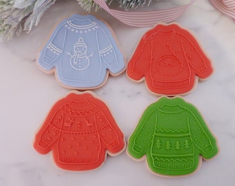 May include: Four sweater-shaped cookies with embossed designs. One is light blue with a snowman, one is orange-red with a Santa face, one is orange-red with reindeer and trees, and one is green with pine trees. Each has a light tan cookie base.