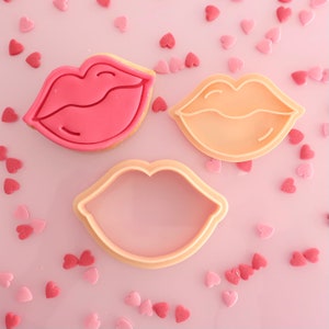 May include: Three lip-shaped items on a pink surface scattered with heart-shaped sprinkles. One is a pink frosted biscuit, another is a peach-coloured biscuit, and the third is a peach-coloured biscuit cutter. The biscuit cutter is made of plastic.