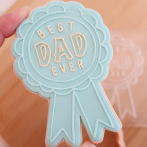 May include: A light blue cookie in the shape of a ribbon with the words "BEST DAD EVER" in light brown icing. The cookie has a scalloped edge and ribbon tails. A second, transparent cookie is visible in the background.