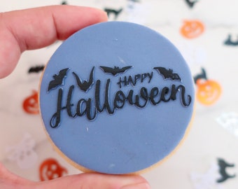 Puede incluir: Una galleta redonda de Halloween con una cobertura azul claro. La galleta está decorada con murciélagos negros y las palabras "Happy Halloween" en una fuente de escritura. La galleta es sostenida por una mano.