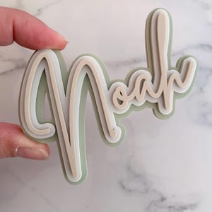 May include: A layered, three-dimensional name cutout spelling "Noah" in a cursive font. The layers are in shades of green, white, and beige. The cutout is held by a hand.