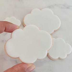 May include: A hand holding a white cloud-shaped biscuit with a light brown border. Several other cloud-shaped biscuits are visible, all with a smooth, white surface and a light brown edge. The biscuits are arranged on a light-coloured surface.