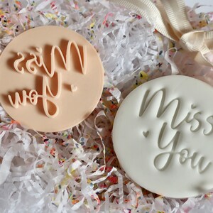 May include: Two circular stamps, one beige and one white, each bearing the words "Miss You" in a cursive script. They rest on a bed of colourful shredded paper, with a cream ribbon visible.