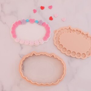 May include: Three cookie cutters in the shape of a bracelet, with a scalloped edge. One is a bracelet with pink and purple star and heart beads. The other two are the same shape, one with a scalloped edge and the other with a smooth edge. Small heart shapes are scattered around.