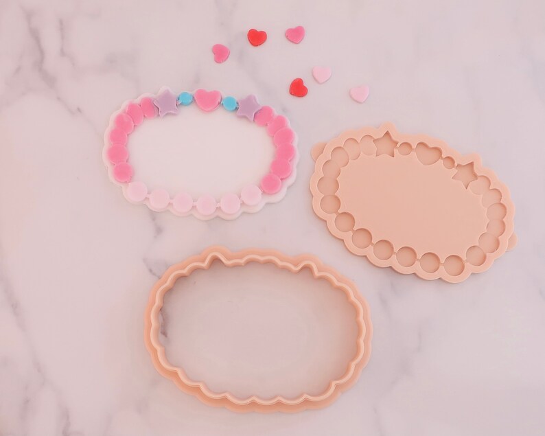 May include: Three cookie cutters in the shape of a bracelet, with a scalloped edge. One is a bracelet with pink and purple star and heart beads. The other two are the same shape, one with a scalloped edge and the other with a smooth edge. Small heart shapes are scattered around.