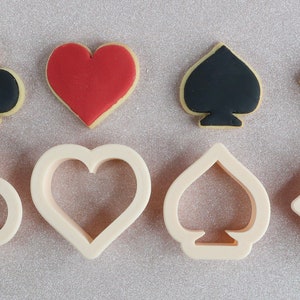 May include: Image shows four playing card suit-shaped biscuits: club, heart, spade, and diamond. Each biscuit has a light-coloured base with a coloured suit symbol. Below are the corresponding biscuit cutters.