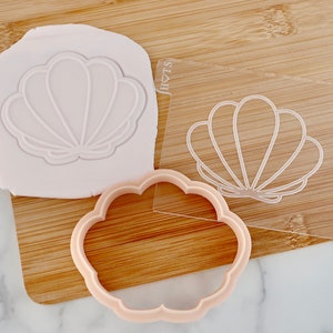 May include: A seashell-shaped cookie cutter and a clear acrylic stamp with the same design are displayed on a wooden board. A piece of pink clay shows the seashell imprint. The cutter is light pink.