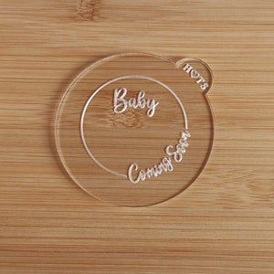 K&ouml;nnte beinhalten: Eine transparente Babyank&uuml;ndigung aus Acryl mit den Worten "Baby Coming Soon" in Schreibschrift. Die Ank&uuml;ndigung ist kreisf&ouml;rmig mit einem herzf&ouml;rmigen Detail und dem Wort "HOTS". Der Hintergrund ist eine hellbraune Holzoberfl&auml;che.