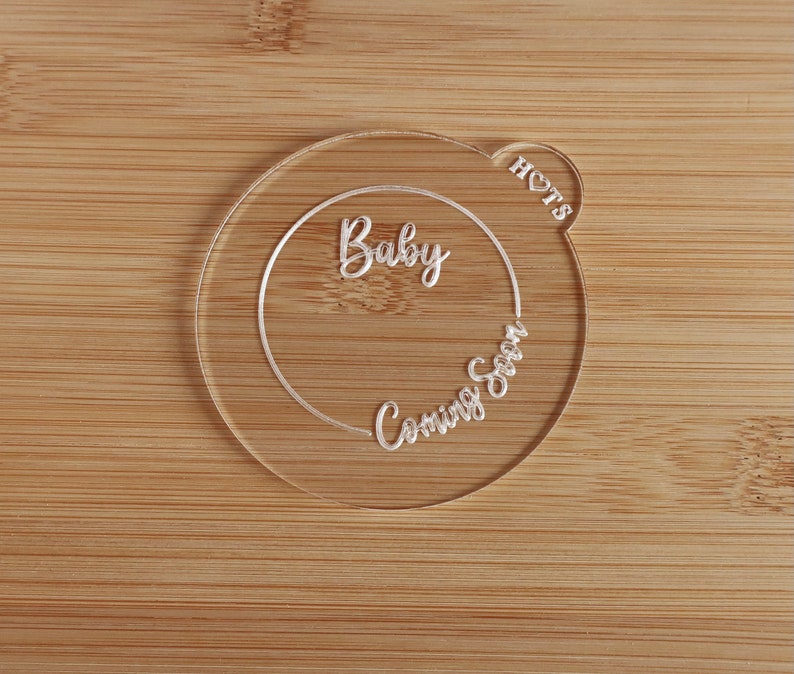 K&ouml;nnte beinhalten: Eine transparente Babyank&uuml;ndigung aus Acryl mit den Worten "Baby Coming Soon" in Schreibschrift. Die Ank&uuml;ndigung ist kreisf&ouml;rmig mit einem herzf&ouml;rmigen Detail und dem Wort "HOTS". Der Hintergrund ist eine hellbraune Holzoberfl&auml;che.