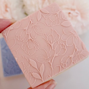 May include: A square, light pink cookie stamp with a floral design. The stamp features an embossed pattern of flowers and leaves. A second, blue stamp is visible in the background. The cookie stamp is held by a person.
