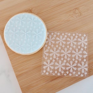 May include: A round cookie with a light blue frosting, decorated with white snowflake patterns. A clear, square stencil with the same snowflake design is next to the cookie. The items are on a wooden surface.
