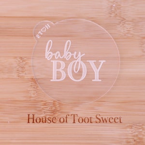 May include: A clear, circular acrylic cake topper with the words "baby BOY" in a delicate script font. The text "House of Toot Sweet" is engraved at the bottom. The topper is placed on a light-colored wooden surface.