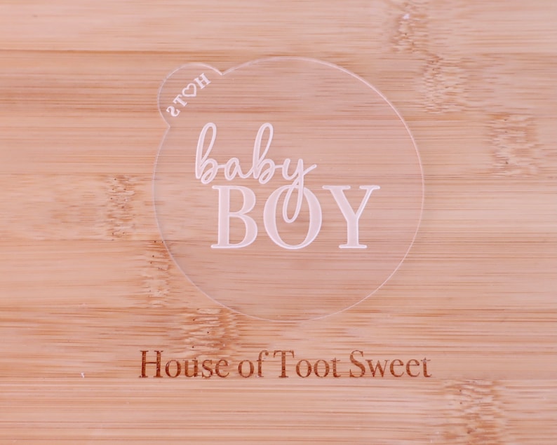 May include: A clear, circular acrylic cake topper with the words "baby BOY" in a delicate script font. The text "House of Toot Sweet" is engraved at the bottom. The topper is placed on a light-colored wooden surface.