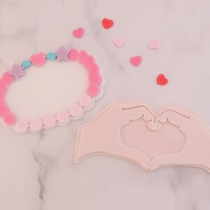 May include: A pink and white bracelet with star and heart-shaped beads, and a hand-shaped design forming a heart. Scattered around are small heart-shaped decorations in shades of pink and red.