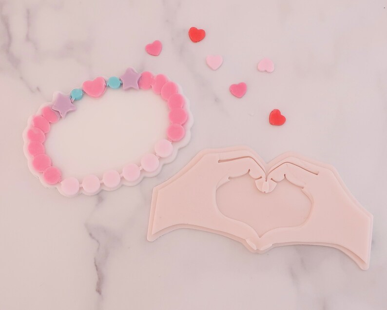 May include: A pink and white bracelet with star and heart-shaped beads, and a hand-shaped design forming a heart. Scattered around are small heart-shaped decorations in shades of pink and red.