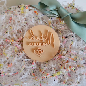 Puede incluir: Un sello redondo de madera clara con las palabras "Nihal Mubarak" en escritura cursiva y un dise&ntilde;o de coraz&oacute;n. El sello est&aacute; en papel triturado blanco con acentos florales coloridos, atado con una cinta de raso verde claro.