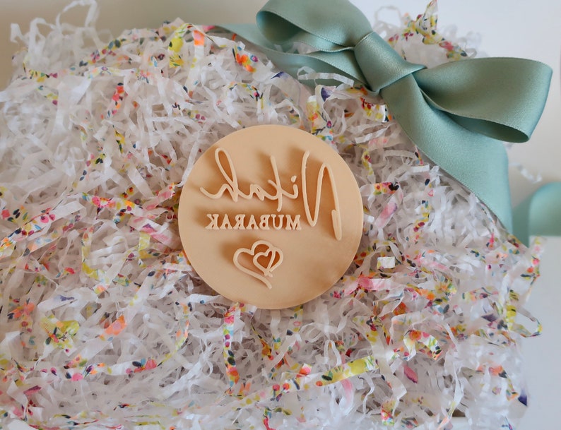 Puede incluir: Un sello redondo de madera clara con las palabras "Nihal Mubarak" en escritura cursiva y un dise&ntilde;o de coraz&oacute;n. El sello est&aacute; en papel triturado blanco con acentos florales coloridos, atado con una cinta de raso verde claro.