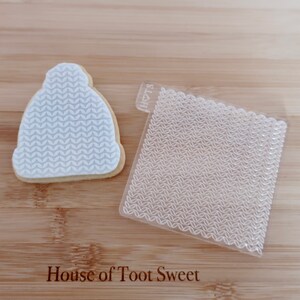 May include: A white biscuit shaped like a beanie with a textured knit pattern and a clear square acrylic embosser featuring the same knit design and the text HTS with a heart symbol, used for creating impressions on baked goods.