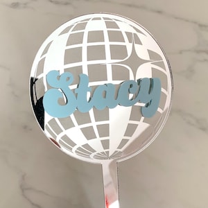May include: A circular cake topper featuring a reflective silver mirror finish with a grid pattern resembling a disco ball. The name Stacy is written across the centre in a light blue, cursive, three-dimensional font with a star accent.