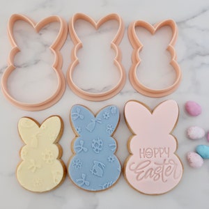 May include: Pink bunny-shaped cookie cutters and decorated cookies with floral and Easter egg designs. One cookie has the text "Happy Easter". Pastel-colored Easter candies are scattered around the cookies.
