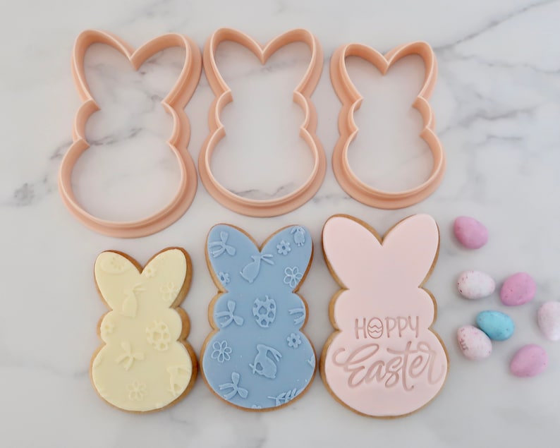 May include: Pink bunny-shaped cookie cutters and decorated cookies with floral and Easter egg designs. One cookie has the text "Happy Easter". Pastel-colored Easter candies are scattered around the cookies.