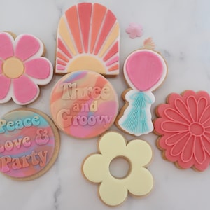 May include: A selection of decorated biscuits in various shapes and designs. The biscuits feature a flower, a sunburst, a balloon, and a daisy. Some biscuits have text that reads "Peace Love & Party" and "Three and Groovy". The biscuits are decorated with pink, orange, yellow, and blue icing.