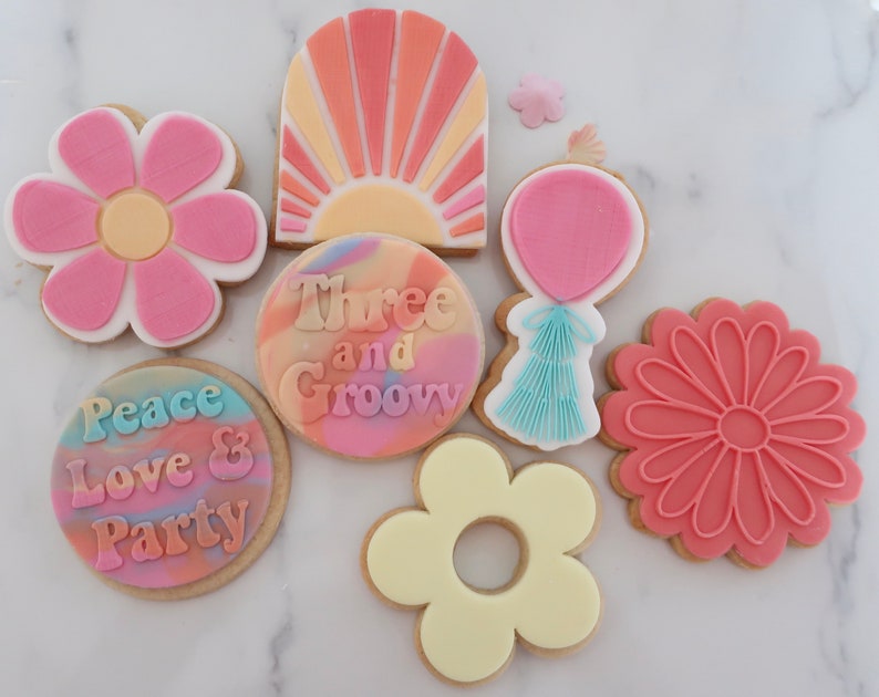 May include: A selection of decorated biscuits in various shapes and designs. The biscuits feature a flower, a sunburst, a balloon, and a daisy. Some biscuits have text that reads "Peace Love & Party" and "Three and Groovy". The biscuits are decorated with pink, orange, yellow, and blue icing.