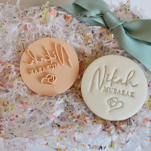 Puede incluir: Dos sellos redondos para galletas, uno beige y otro blanco, con las palabras "Nikah Mubarak" y un dise&ntilde;o de coraz&oacute;n. Se presentan en una caja de regalo con papel floral y una cinta color turquesa.
