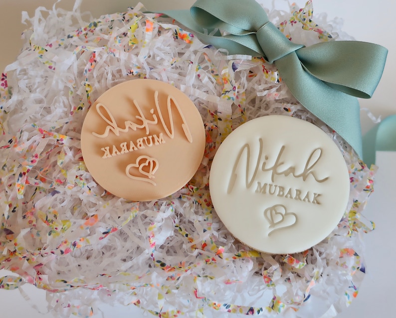 Puede incluir: Dos sellos redondos para galletas, uno beige y otro blanco, con las palabras "Nikah Mubarak" y un dise&ntilde;o de coraz&oacute;n. Se presentan en una caja de regalo con papel floral y una cinta color turquesa.