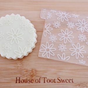 May include: A stack of white cookies with a daisy pattern pressed into the top, next to a clear acrylic stamp with a daisy design. The text "House of Toot Sweet" is printed at the bottom of the image.