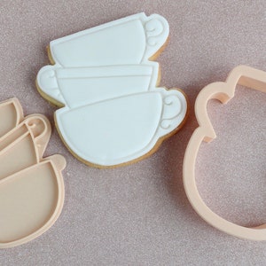 May include: A collection of teacup-shaped biscuits, iced in white, alongside a matching cookie cutter. The biscuits are outlined in light brown. The cutter and biscuits are on a pink, glittery surface. The biscuits are decorated with a light brown outline.