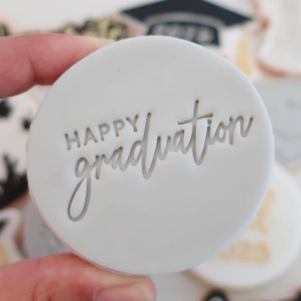 May include: A white, round cookie with the words "HAPPY graduation" imprinted in a cursive font. The cookie is held in a hand, with other graduation-themed cookies visible in the background.