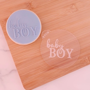 May include: A light blue cookie with the words "baby BOY" imprinted on it, next to a clear plastic stamp with the same text. The cookie sits on a wooden cutting board.