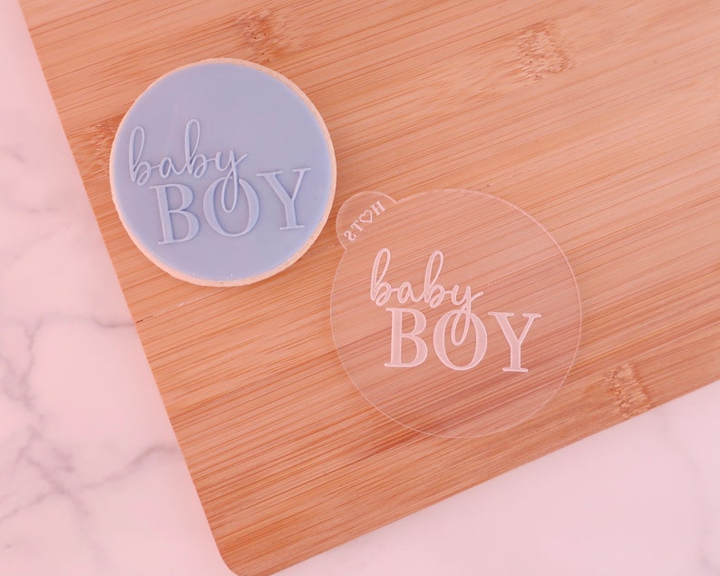 May include: A light blue cookie with the words "baby BOY" imprinted on it, next to a clear plastic stamp with the same text. The cookie sits on a wooden cutting board.