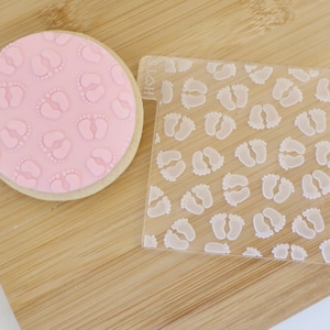 May include: A round biscuit with pink icing decorated with baby footprints. Beside it is a clear acrylic stamp, also with a footprint pattern. The items are displayed on a light wooden surface.