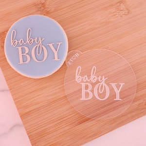 May include: A light blue frosted cookie with the words "baby BOY" in a cursive font. A clear, circular acrylic piece with the same text is next to the cookie. The items are on a wooden surface.