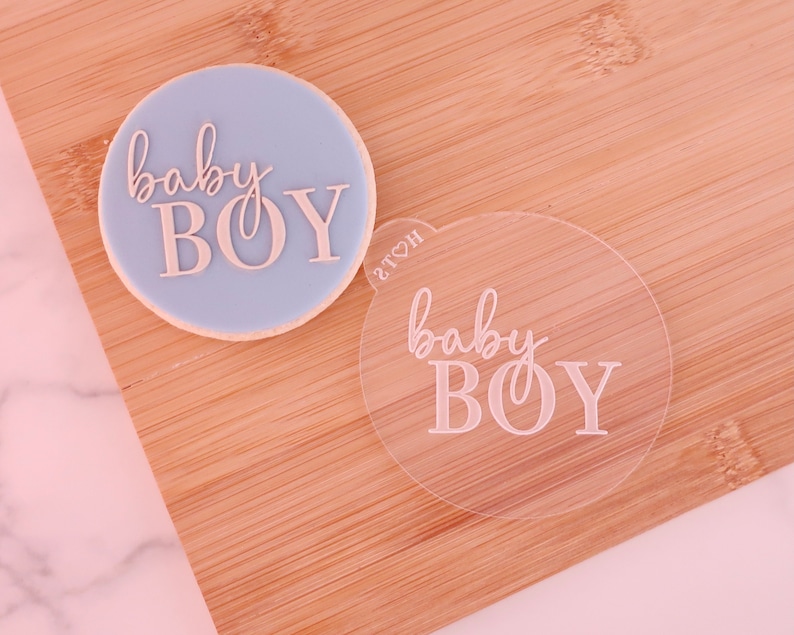 May include: A light blue frosted cookie with the words "baby BOY" in a cursive font. A clear, circular acrylic piece with the same text is next to the cookie. The items are on a wooden surface.