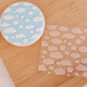 May include: A round biscuit with light blue icing and a white cloud design. A clear plastic stencil with a cloud pattern is next to the biscuit. The biscuit is on a wooden chopping board.