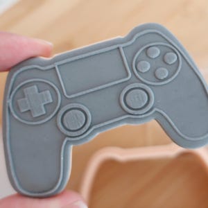 May include: A gray, plastic cookie cutter in the shape of a video game controller. The controller has a directional pad, buttons, and a central screen area. The cutter is held by a person's fingers.