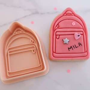 May include: A pink backpack-shaped cookie cutter and a decorated cookie. The cookie is pink with a backpack design, a star, a heart, and the name "MILA". The cutter is made of light pink plastic.