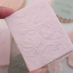 May include: A light pink square biscuit with a floral pattern. The design features embossed flowers and leaves. Other biscuits with similar designs are in the background.